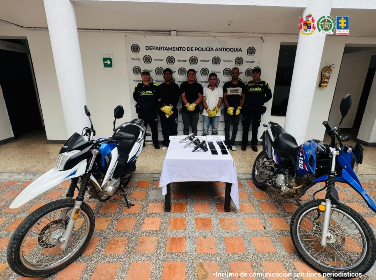 En la imagen aparecen los procesados en medio de dso uniformados de la Policía Nacional. Frente a ellos hay dos motocicletas y una mesa con tres pistolas y varios celulares.
