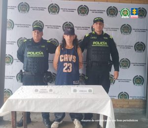En la foto aparece el capturado de frente y custodiado por dos uniformados de la Policía Nacional. En la foto también aparece un backing de la Policía Nacional y una mesa.
