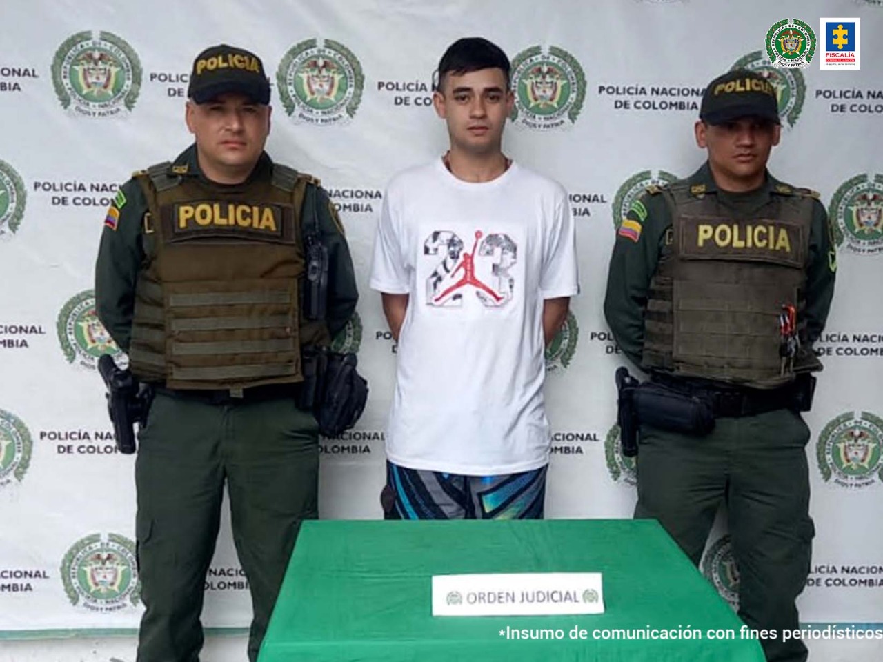 Se observa al centro el capturado y a cada uno de sus costados un funcionario de Policía Nacional. Al fondo un pendón de Policía Nacional. Al frente una mesa con el cartel 'ORDEN JUDICIAL.