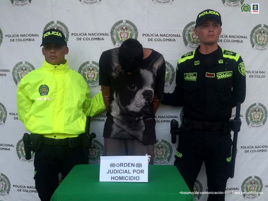 Se observa al capturado y en cada costado un funcionario de Policía Nacional. Atrás un pendón de Policía Naciones y adelante una meda con el aviso: orden judicial por homicidio