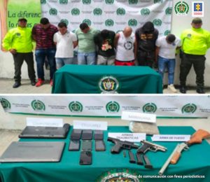 En la imagen se ven 7 personas detenidas entre dos Policías. A su vez, una mesa con celulares, armas de fuego, computadores y munición.
