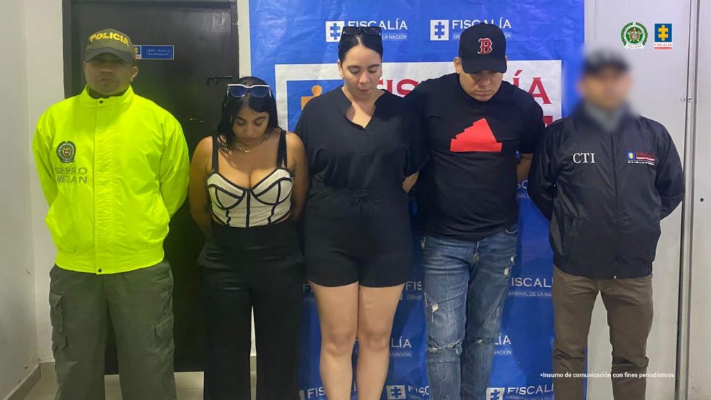 Tres de los capturados se encuentran de frente y custodiados por un servidor del CTI y la Policía Nacional. Detrás se observa un backing de la Fiscalía