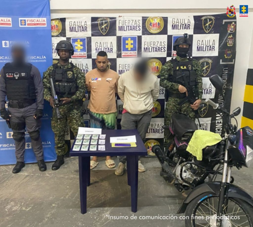 En la fotografía se aprecia de pie y con los brazos atrás a Bladimir Padilla Marín, alias Blacho. Viste camiseta color naranja pálido, bermuda estampada con colores negro, amarillo y pantuflas. A su extremo derecho hay un servidor del CTI de la Fiscalía y un uniformado del Ejército. A su izquierda hay un uniformado del Ejército. Delante de ellos hay una mesa cuadrada, oscura, y sobre ella hay dinero en efectivo y un teléfono celular. Al extremo izquierdo del procesado hay una motocicleta. Detrás de ellos hay dos pendones institucionales de la Fiscalía y otro con logos del Gaula Ejército y de la Fiscalía.