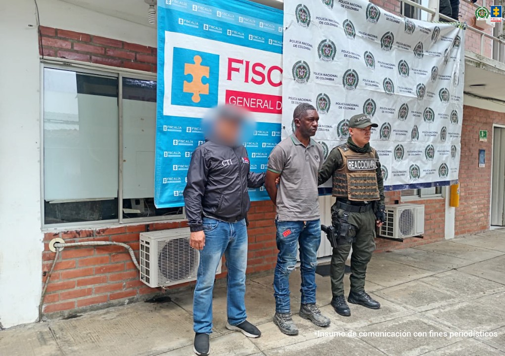 En la fotografía se aprecia de pie y con los brazos atrás a Bleyner Adrián Mejía Brand.Viste con camisa de cuello, mangas cortas, de color gris con ribetes verdes en el borde del cuello y de las mangas; jean azul desgastado y zapatos negros. Al extremo derecho de él hay un servidor del CTI de la Fiscalía y al izquierdo un uniformado de la Policía Nacional. Detrás de ellos hay dos pendones institucionales de la Fiscalía y la Policía.