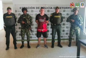 capturado de frente esposado manos atrás custodiado por policías y militares. Detrás banner de la POlicía Nacional.