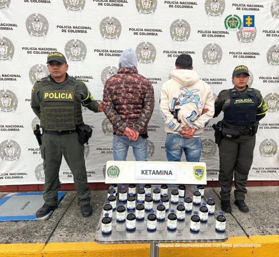 En la fotografía aparecen de pie, de espaldas a la cámara, con los brazos atrás y esposados, Brayan Alexander Coral Betancourth y Juan David Serrano Álvarez. Uno viste con chaqueta estampada de capucha de color gris y jean azul claro. El otro vista con buso de color beage con estampado atrás, jean azul claro desgastado. En sus extremos hay uniformados de la Policía Nacional. En la parte delantera, sobre el suelo hay varios frascos con etiqueta y un letrero que dice ‘Ketamina’. En la parte posterior hay un pendón institucional de la Policía.