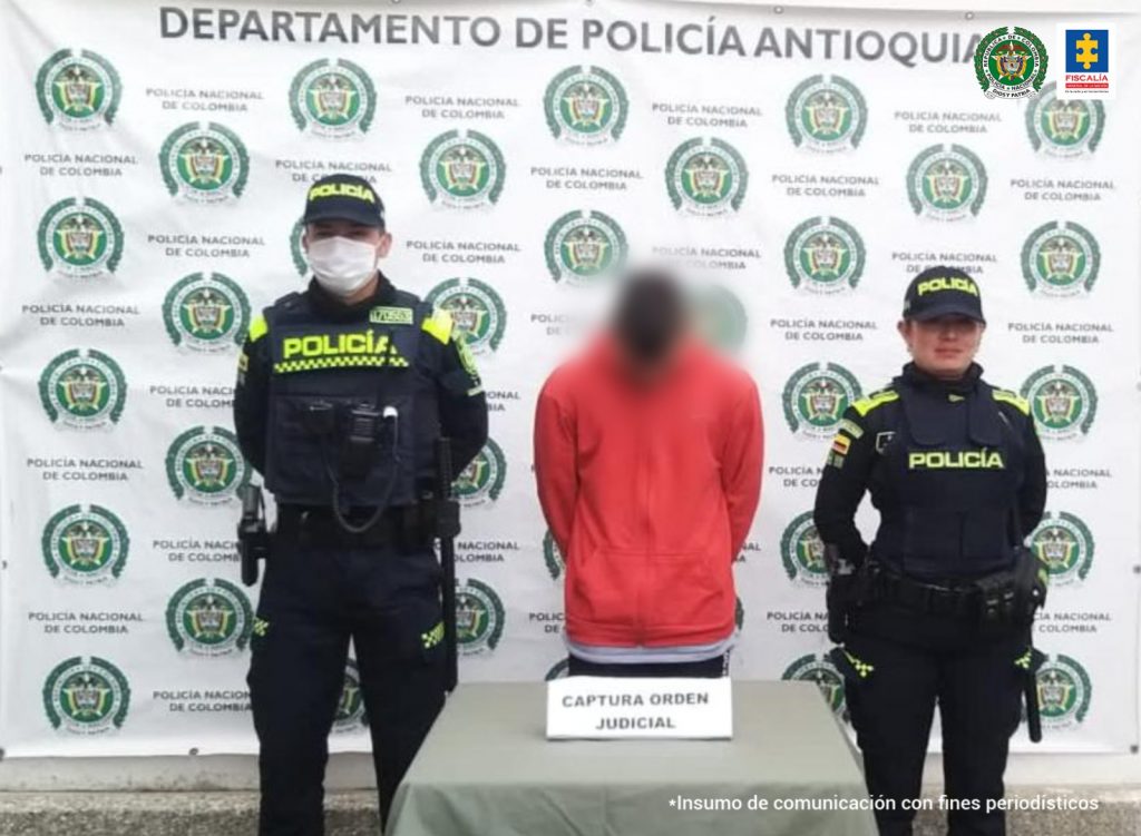 En el centro se observa el capturado y a cada lado un funcionario de Policía nacional. Al fondo un pendón de Policía Nacional.