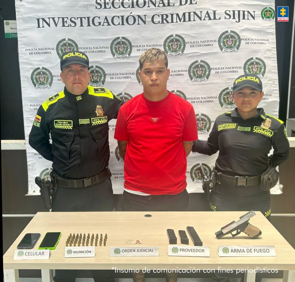 En la imagen se observa al capturado junto a dos uniformados de la Policía Nacional. Frente a ellos una mesa con los elementos incautados. Detrás de ellos el banner que identifica a la Policía Nacional.