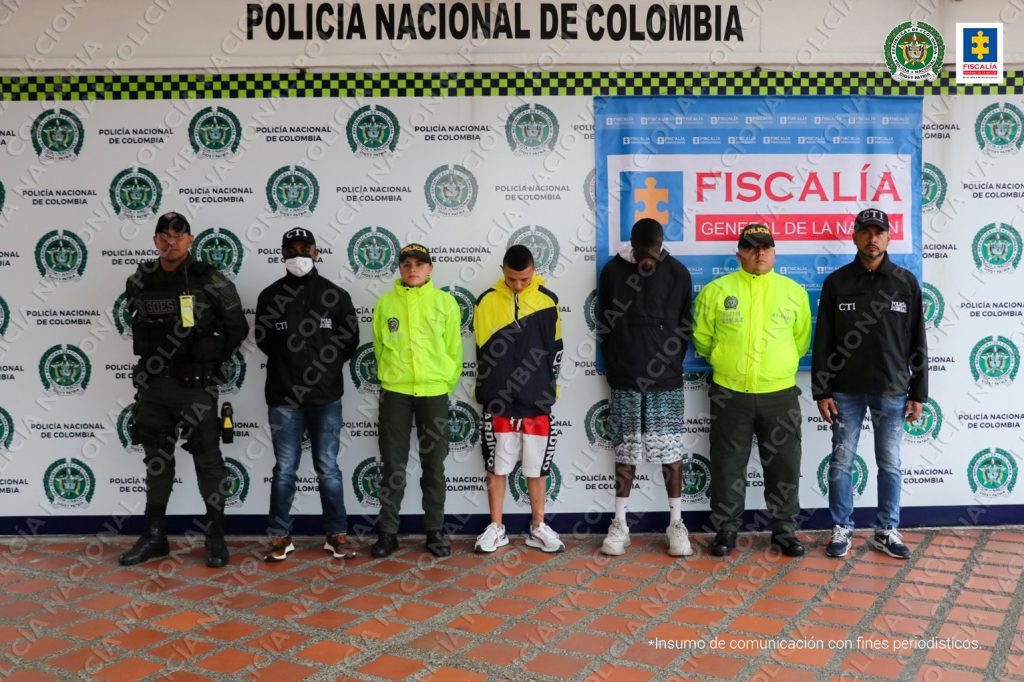 Se observan en el centro dos capturados, a un lado de la fila un funcionario de la Policía, uno del CTI y uno del Gaula. Al otro lado un funcionario de la Policía y uno del CTI. Atrás un pendón de la Policía y otro de la Fiscalía