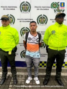 En la foto aparece, de pie y con los brazos atrás, Bryan Andrés Gutiérrez Guerrero. Esta persona viste camiseta de manga corta, blanca, naranja y azul claro; así como estampados negros, jean azul y tenis blancos. A lado y lado de él hay uniformados de la Policía Nacional. Detrás de ellos hay un pendón institucional de la Policía Nacional.