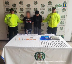 En la fotografía se aprecia de pie y los brazos atrás a Bryan Esteban Molano Ospina. Viste camiseta de anga corta de color negro y bermuda de color gris oscuro. A sus extremos hay uniformados de la Policía Nacional. Delante de ellos hay una mesa con mantel blanco que tiene bordado el logo de la Policía y sobre él hay municipio, cigarrillos, una bolsa plástica con contenido vegetal un contenedor abierto, de color azul, y en su interior hay una bolsa plástica. Asimismo, tres letreros que dicen: ‘Munición’, ‘Elementos de empaque’ y ‘Marihuana’. Detrás hay un pendón institucional de la Policía.