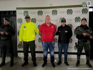 En la foto aparece, de pie y con los brazos atrás, Bryan Steven Soto Mosquera, alias ‘El Mono Bryan’. Esta persona viste camisa roja de manga corta y cuello; así como jean azul y zapatos oscuros. Al lado derecho de él hay uniformados de la Policía Nacional y a la izquierda hay un servidor del CTI de la Fiscalía y un uniformado de la Policía. Detrás de ellos hay un pendón institucional de la Policía Nacional.