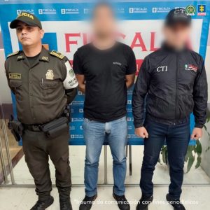 En la fotografía aparece de pie y con los brazos atrás el hoy imputado. Viste con camiseta de mangas cortas, de color negro; así como jean azul claro y zapatillas negras. En su extremo izquierdo hay un servidor del CTI de la Fiscalía y en el derecho un uniformado de la Policía Nacional. Detrás de ellos hay un pendón institucional de la Fiscalía.