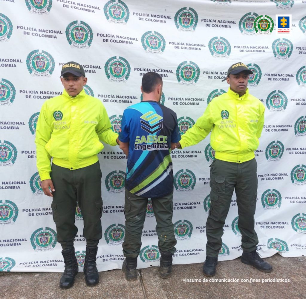 🔼 El capturado aparece de espalda a la cámara y custodiado por dos uniformados de la policía nacional y hay un pendón institucional de fondo.