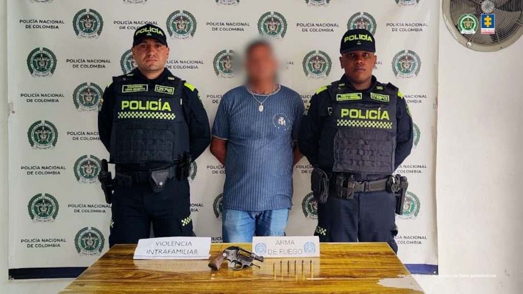 Se observa al capturado, de frente, custodiado a ambos costados por servidores de Policía Nacional. Atrás, un pendón de la Policía Nacional. Adelante, una mesa con el arma y munición incautados, un letrero que dice VIOLENCIA INTRAFAMILIAR y otro, ARMA DE FUEGO