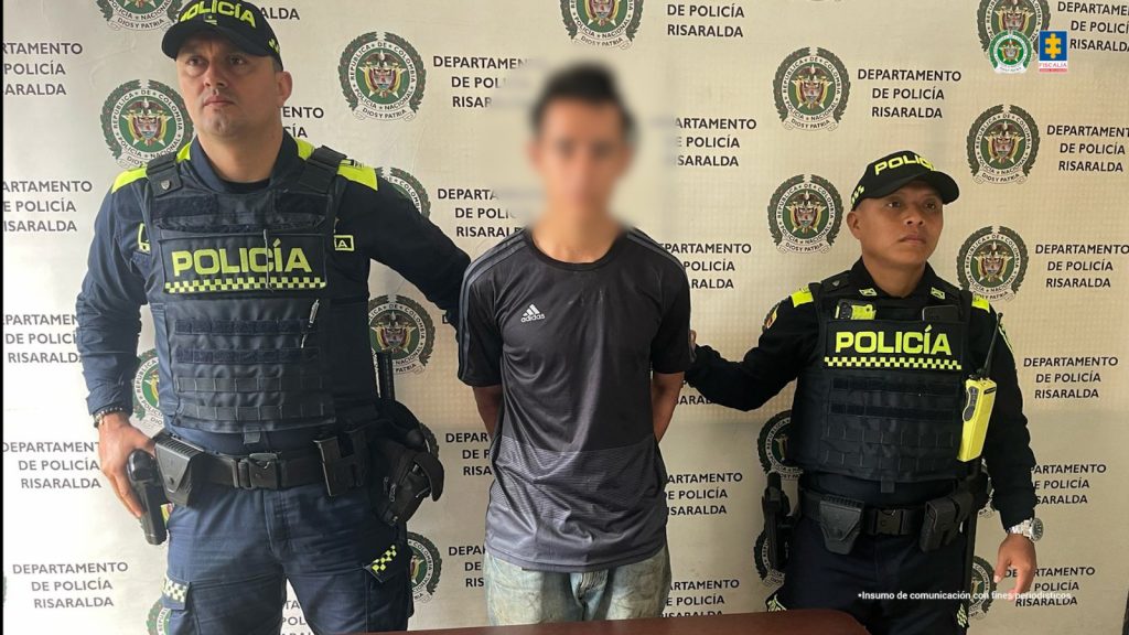 Se observa al capturado, de frente, custodiado a ambos costados por servidores de Policía Nacional. Atrás, un pendón de Policía Nacional