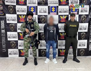 El imputado se encuentra en custodia por un uniformado de la Policía y Ejército Nacional. Detrás hay un backing del Ejército.