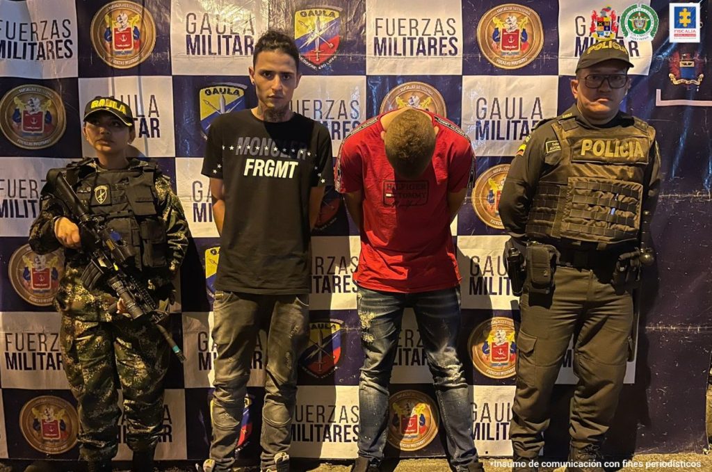 Se observan los dos detenidos. En un costado un funcionario de Ejército nacional y al otro uno de Policía Nacional. Atrás un pendón de Ejército Nacional