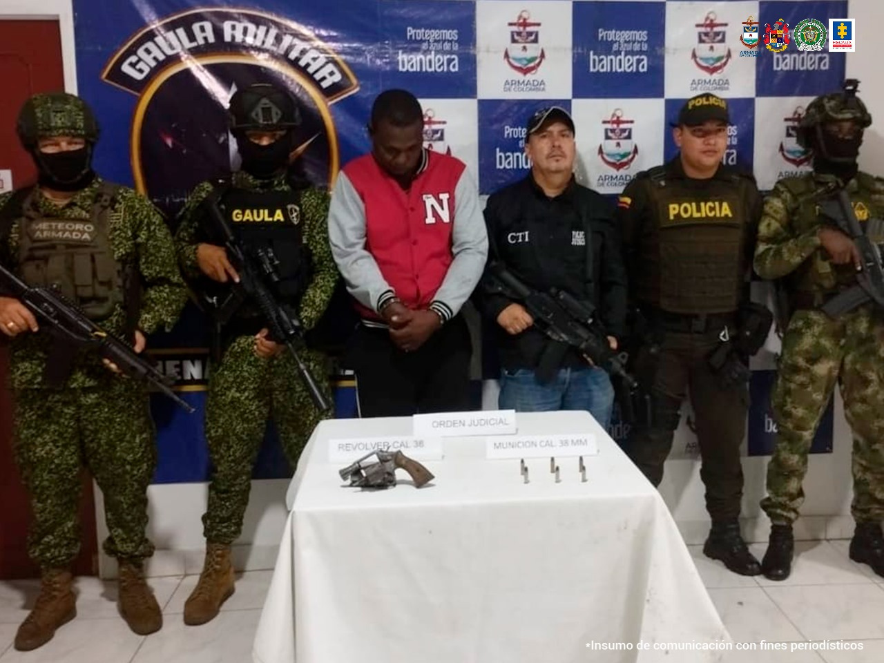 En la fotografía aparece el capturado junto a un servidor del CTI, un agente de la Policía, uniformados del Gaula Militar y la Armada Nacional. En la parte superior está una mesa con un arma de fuego y munición