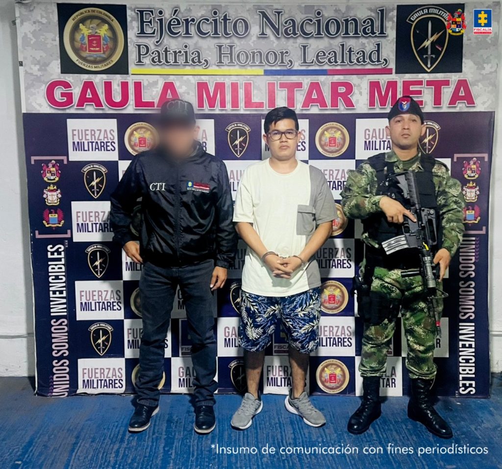 capturado de frente custodiado por investigador del CTI de la Fiscalía y un militar. Detrás banner del grupo Gaula Militar.