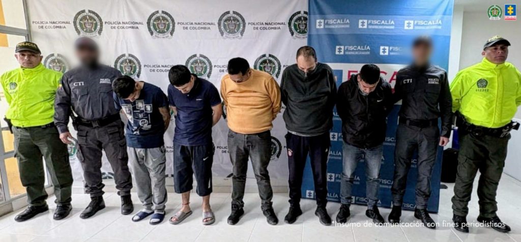 en la fotografia se puede ver a los 5 capturados en medio de dos agentes del CTI de la Fiscalia y dos funcionarios de la policia nacional, detras de ellos se pueden ver los bankings de la policia y la fiscalia