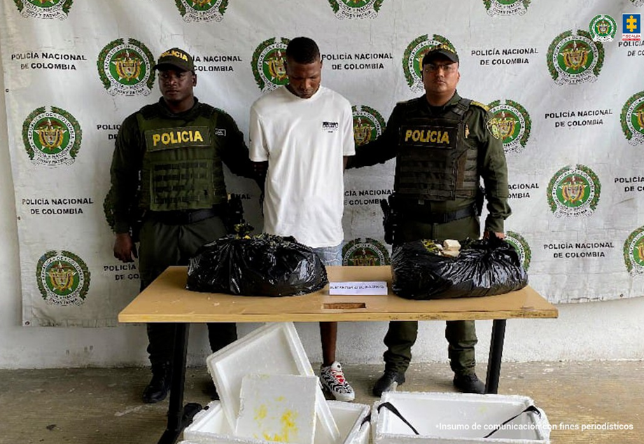 Un hombre con camiseta blanca y manos esposadas es custodiado por dos policías uniformados. Frente a ellos hay una mesa con bolsas negras que contienen paquetes de droga incautados y neveras de icopor abiertas. Al fondo se ve un pendón de la Policía Nacional de Colombia.
