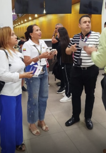 Cuatro personas con credenciales visibles conversan con otra persona; visten prendas en tonos blanco, azul y negro, y sostienen papeles en un espacio con paredes amarillas y pantallas negras. Funcionarios de la Fiscalía orientan a usuarios en el aeropuerto, entregando información sobre canales de atención y acceso a la justicia.