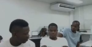 En la imagen aparecen tres hombres de tes oscura vestidos con camisetas blancas. De fondo se ve una ofician con computadores. 