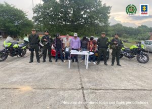capturados de frente custodiados por la Policía, denate mesa con material incautado. Detrás vehículos incutados.