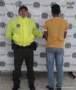 En la foto se ve un hombre de espaldas custodiado por un uniformado de la Policía Nacional, detrás un banner de la Policía.