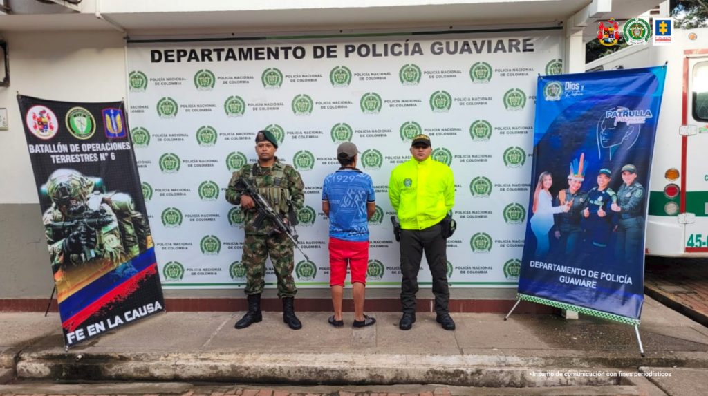 Están los capturados de frente esposados manos atrás, custodiados por dos uniformados de la Policía. Detrás banner de la Policía Nacional.
