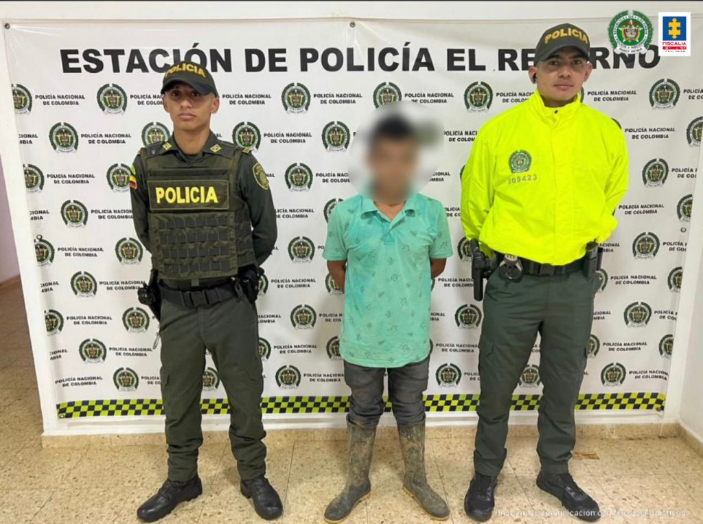 capturado de frente esposado custodiado por uniformados de la Policía, detrás banner de la Policía Nacional.