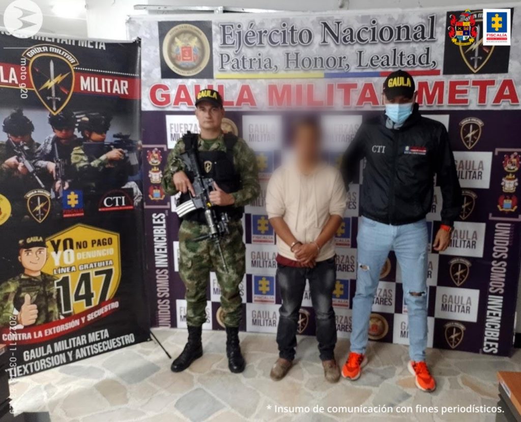 Está el capturado esposado con manos adelante con pantalón y saco, custodiado por investigador de CTI de Fiscalía Gaula y soldad de grupo Gaula Militar detrás banner con logos de Fiscalía y Ejército.