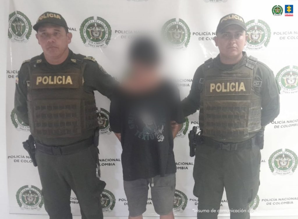 capturado de frente custodiado por dos policías, detrás bannerde la Policía Nacional.