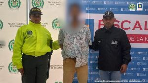 capturado de frente esposado manos atrás, custodiado por un policía y un investigador del CTI de la Fiscalía, detrás banner de Policía Nacional y Fiscalía.