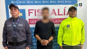 Por delitos sexuales contra una mujer y una menor de edad son asegurados dos hombres en Meta capturado con gorra y gafas, esposado de frente y custodiado por investigador del CTI de la Fiscalía y un policía. Detrás banner de las Fiscalía.