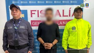 capturado con gorra y gafas, esposado de frente y custodiado por investigador del CTI de la Fiscalía y un policía. Detrás banner de las Fiscalía.