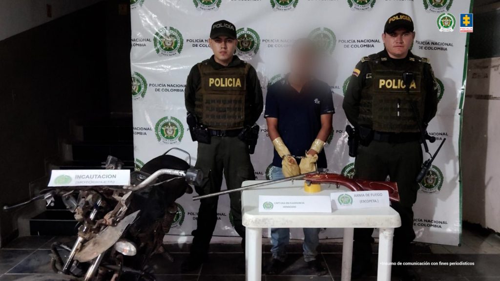 detenido de frente esposado custodiado por dos policías. delante de él mesa con escopeta y motocicleta incautadas.