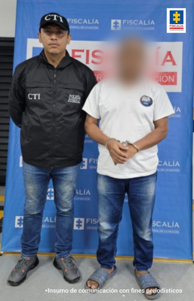 capturado está esposado, manos adelante, custodiado por investigador del CTI de la Fiscalía, detrás banner de la Fiscalía.