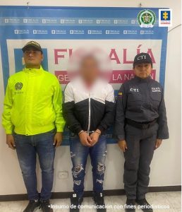 capturado de frente esposado manos adelante, custodiado por un policía y un investigador del CTI de la Fiscalía. Detrás banner de Fiscalía.
