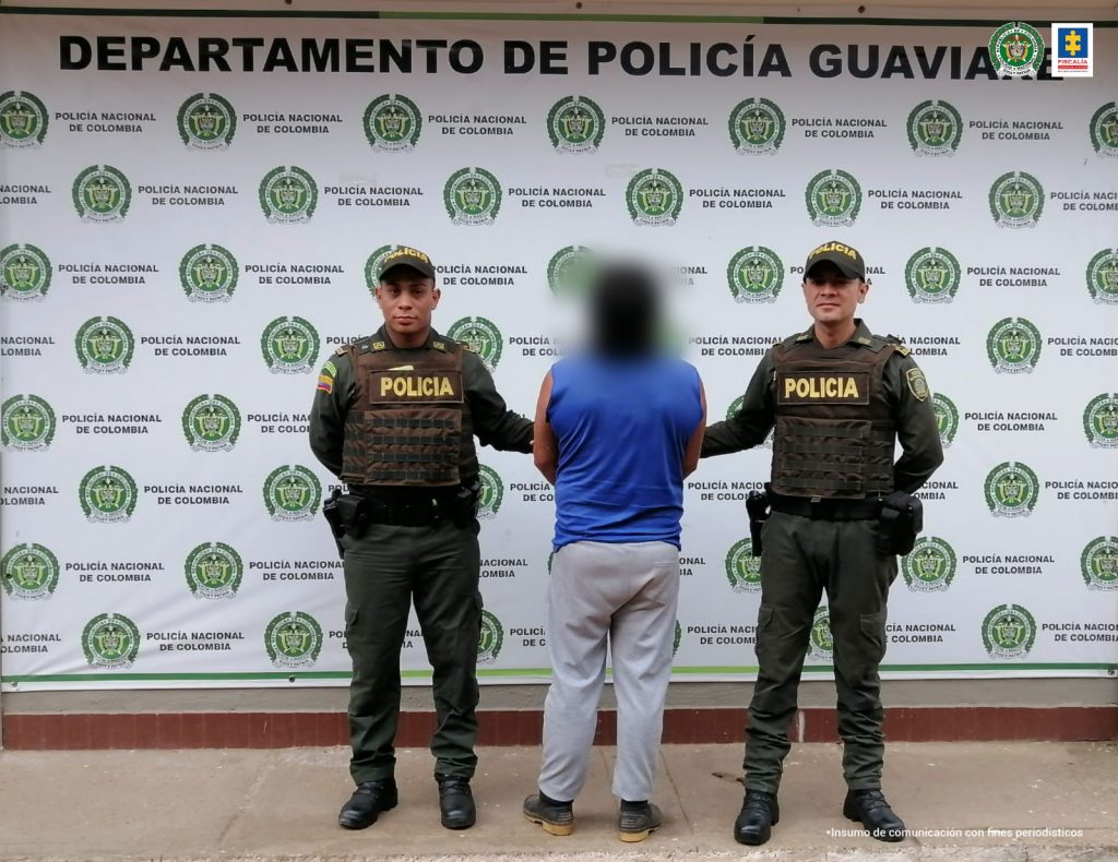 capturado de espaldas custodiado por dos uniformados de la Policía Nacional, detrás banner de la Policía Nacional.
