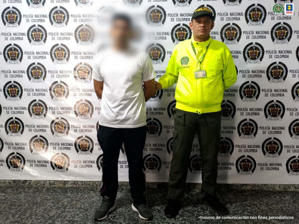 el capturado está esposado con las manos atrás custodiado por un policía uniformado, detrás banner de la Policía Nacional.