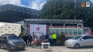 capturados de espaldas, custodiados por investigadores del CTI de la Fiscalía, policías y militares del Ejército Nacional. En torno a los capturados un camión, dos vehículos, una motocicleta y un arma traumática incautados en la diligencia judicial. Detrás banner de Policía y Ejército Nacional.