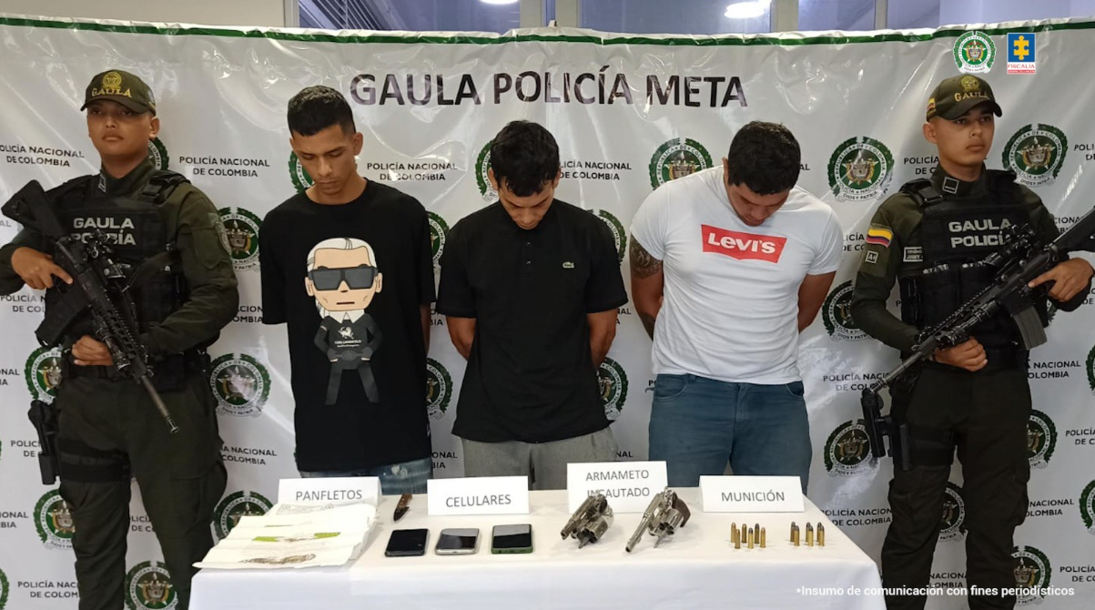 Capturados de frente custodiados por policías. Detrás banner de la Policía Nacional y delante de los capturados mesa con material incautado.