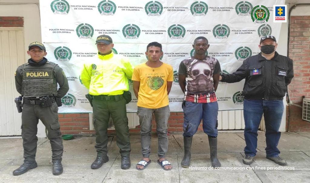 En la fotografía se observan cinco personas, dos de ellas los capturados, dos uniformados de la Policía Nacional y el último un funcionario del CTI. En la parte posterior se encuentra un backing de la Policía Nacional.