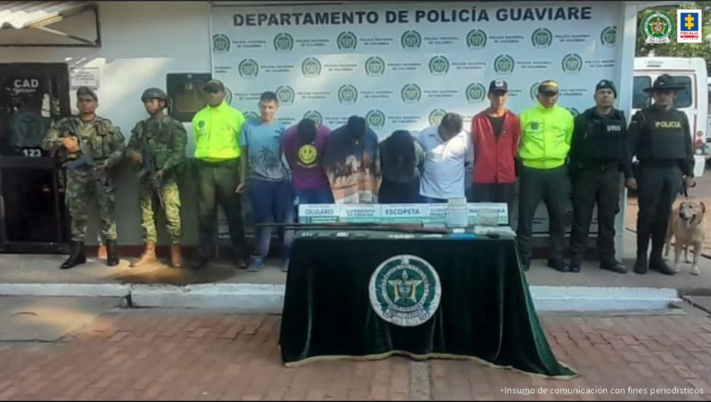 capturados de frentes esposados, custodiados por policías y militares. Delante mes con material incautado, detrás banner de la Policía Nacional.