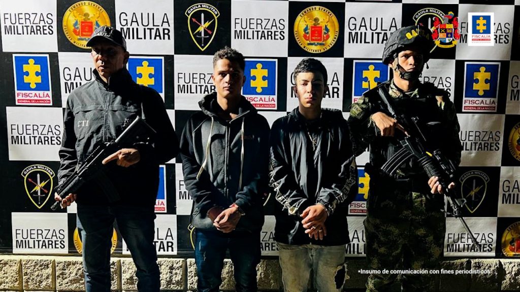 En la imagen están los dos procesados con servidores del CTI y el Gaula del Ejército Nacional y al fondo un pendón con un mosaico de los logos de la Fiscalía y el Ejército.