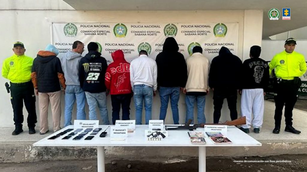 en la imagen se pueden ver los 9 capturados de espaldas en medio de dos funcionarios de la policia. detras de ellos aparece un baking de la policia y en frente de los capturados hay una mesa en donde se pueden ver todos los elementos incautados, entre ellos armas, celulares y libretas