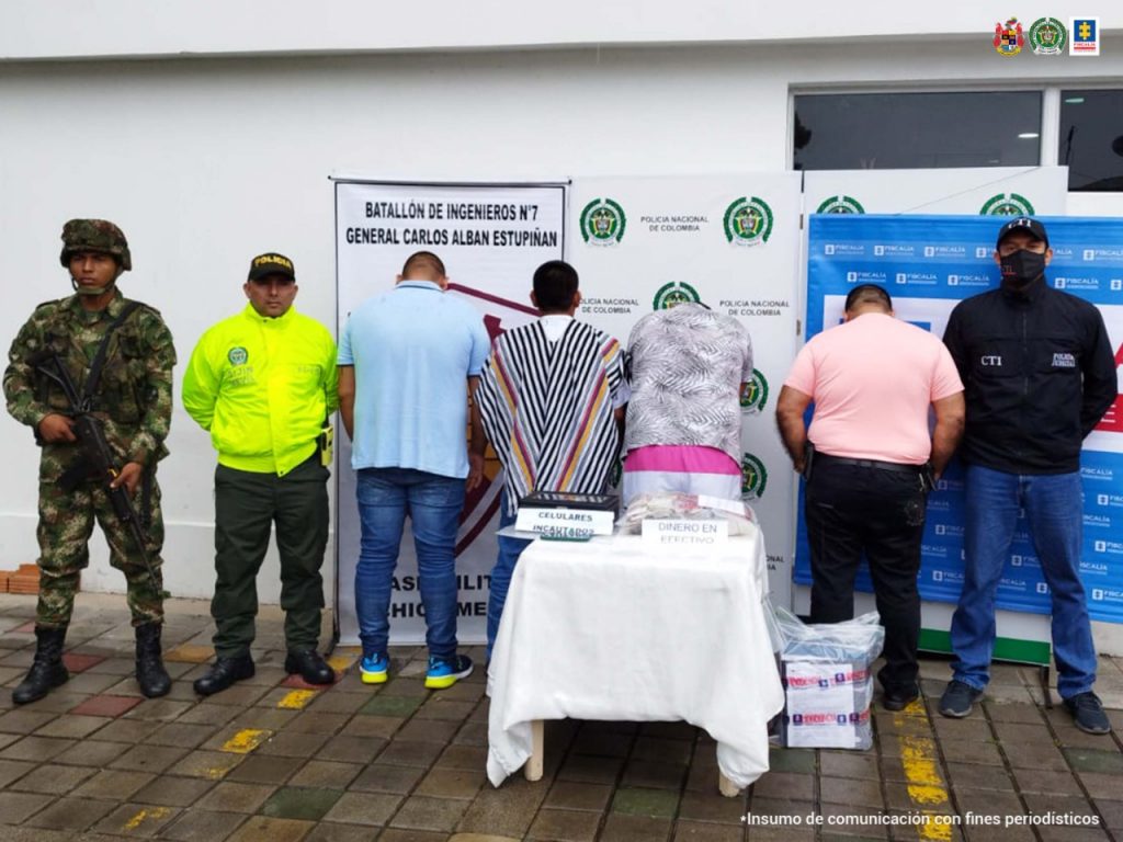En la foto hay cuatro hombres de espaldas, cabeza agachada y manos hacia adelante, custodiados por un investigador del CTI de la Fiscalía y un Militar del Ejército Nacional. Detrás de ellos banner de Ejército, Policía y Fiscalía. Delante de los capturados una pequeña mesa con material incautado.