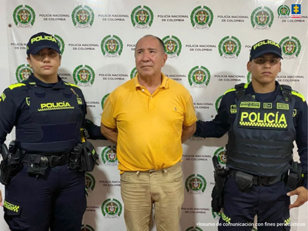 En la fotografía se aprecia de pie y los brazos atrás a Carlos Alberto Correa Martínez. Viste con camisa de color amarillo mostaza y pantalón caqui. A sus extremos hay uniformados de la Policía Nacional. Detrás de ellos hay un pendón institucional de la Policía.
